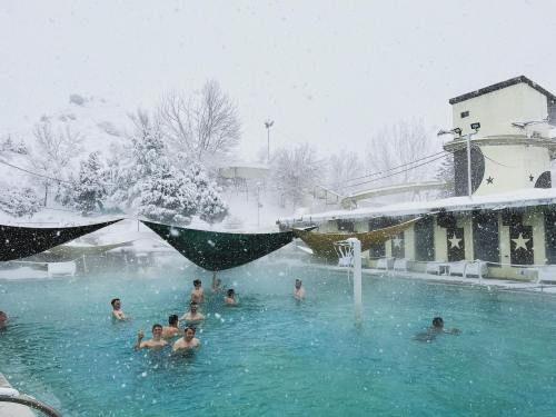 People enjoying a warm pool surrounded by snow, with snowflakes falling and trees in the background.