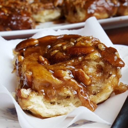 A close-up of a sticky cinnamon roll topped with caramel and nuts, served on a white paper.