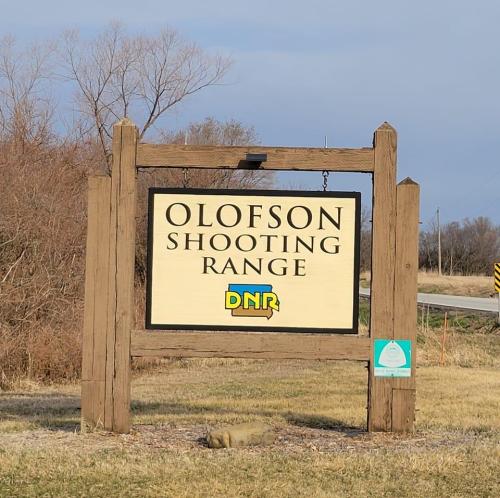 Sign for Olofson Shooting Range, featuring the DNIR logo, set against a clear sky and grassy area.