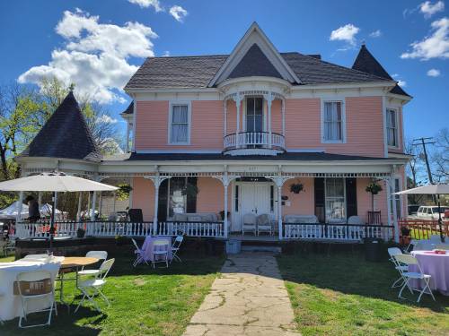 A pink Victorian house with a porch, surrounded by tables and chairs on a sunny day with fluffy clouds.