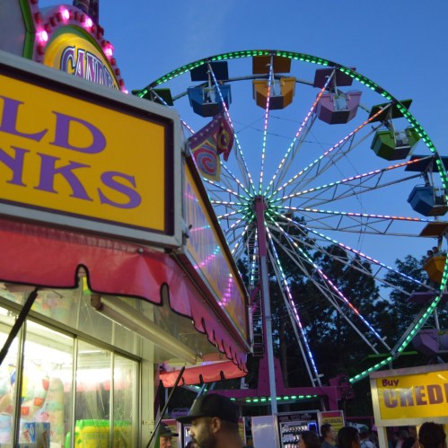 A colorful carnival scene featuring a Ferris wheel and a food stand with bright signage at dusk.