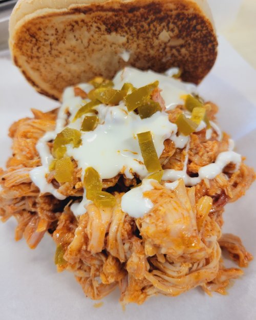 A pulled chicken sandwich topped with sauce, pickles, and drizzled with white sauce, served on a toasted bun.