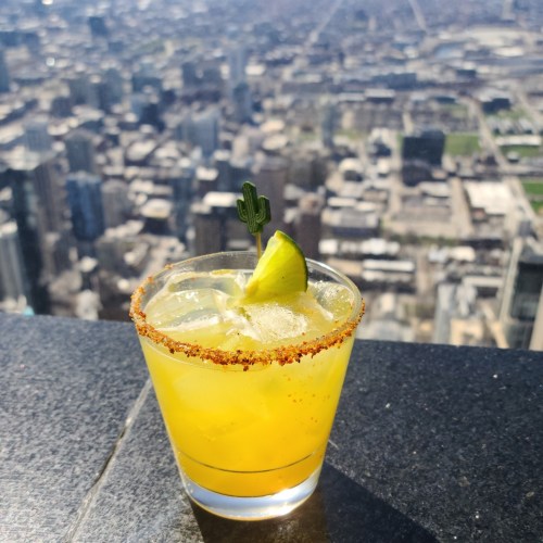 A refreshing yellow cocktail with a lime wedge and cactus garnish, set against a city skyline backdrop.