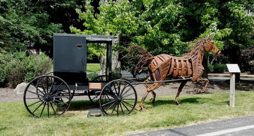 A metal sculpture of a horse pulling a vintage carriage, set in a grassy area with trees in the background.