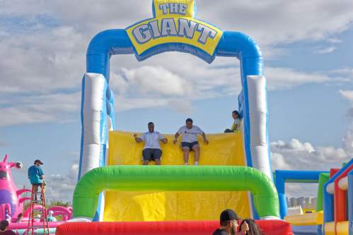 Two people slide down a large inflatable slide labeled "The Giant" at a fun outdoor event. Colorful inflatables are in the background.
