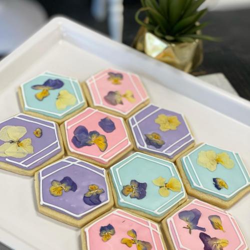 A tray of hexagon-shaped cookies decorated with pastel icing and edible flowers in shades of pink, blue, and purple.