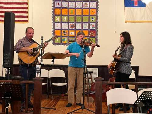 Three musicians perform on stage: one with a guitar, one with a violin, and another with a mandolin, in a community hall.