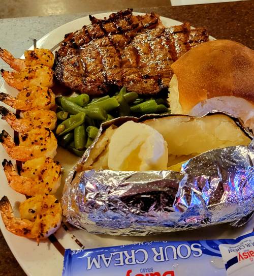 A plate featuring grilled shrimp, steak, green beans, a baked potato with sour cream, and a dinner roll.