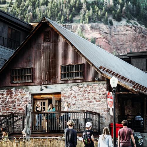 A rustic building with a metal roof, surrounded by trees and mountains, with people on the porch and walking nearby.