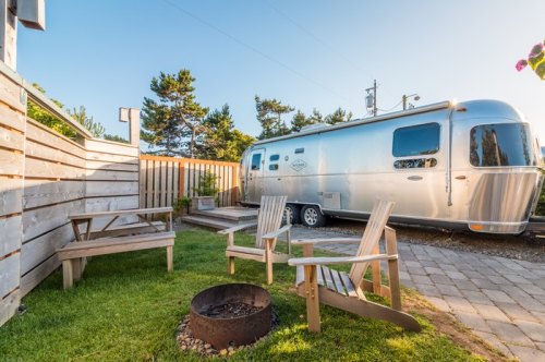 A shiny Airstream trailer parked beside a cozy fire pit and wooden chairs in a grassy outdoor space.