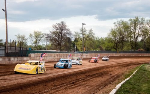 Five race cars compete on a dirt track, with trees and cloudy skies in the background.