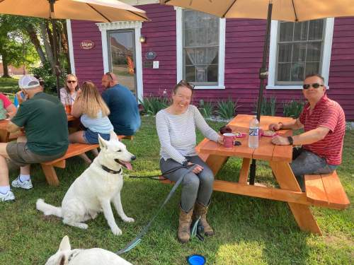 A group of people sitting at picnic tables outside, with two dogs and a purple building in the background.