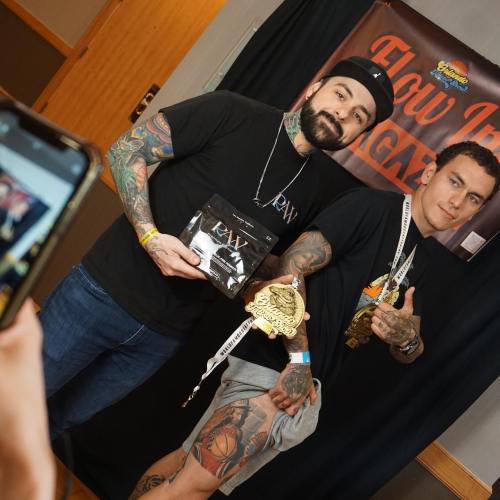 Two tattooed men pose with medals and trophies in front of a black backdrop, smiling at the camera.