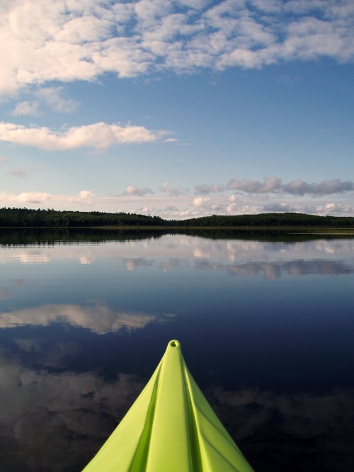 A bright green kayak glides over a calm lake, reflecting clouds and trees under a blue sky.