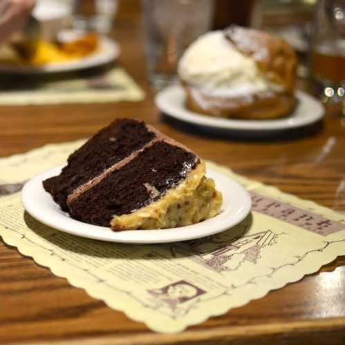 A slice of chocolate cake with a coconut topping on a white plate, set on a wooden table.