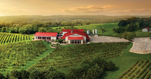 A scenic vineyard with a red-roofed building surrounded by lush green fields and rolling hills at sunset.