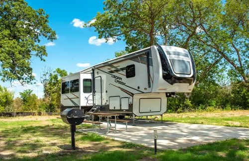 A modern RV parked on a concrete pad surrounded by trees and blue skies. A picnic table and grill are nearby.