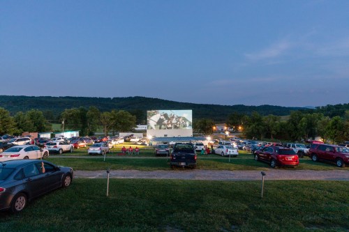 A drive-in movie theater at dusk, with cars parked on a grassy lot and a large screen displaying a film.