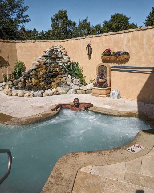 A man relaxes in a bubbling hot tub surrounded by a stone waterfall and lush greenery.