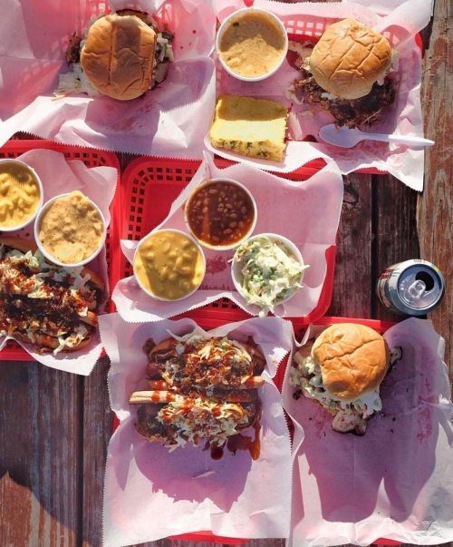 A spread of barbecue dishes including sandwiches, sides, and drinks on red trays, set on a wooden table.