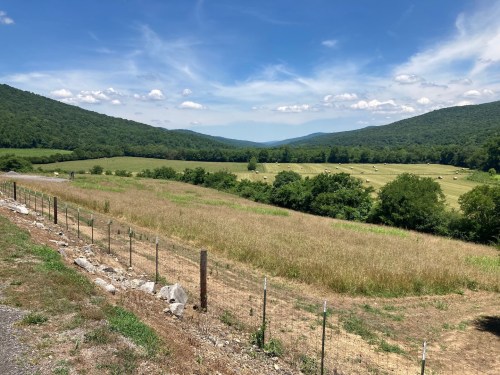 A scenic view of rolling hills and fields under a blue sky with fluffy clouds, featuring hay bales in the distance.