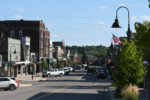 A small town street lined with shops, parked cars, and American flags under a clear blue sky.