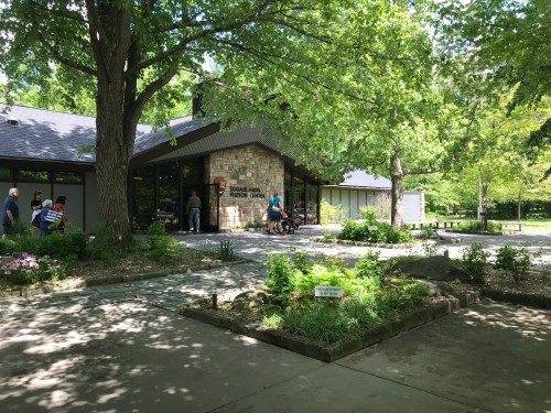 A stone building surrounded by trees, with people entering and walking outside, showcasing a nature center.