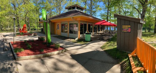 A colorful building with outdoor seating, surrounded by trees and a playground, on a sunny day by the water.
