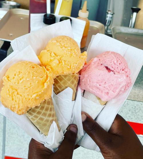 Three ice cream cones in hand: one orange, one pink, and one yellow, wrapped in white paper.