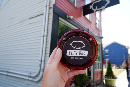 A hand holds a red buzzer for Eli's BBQ, with the restaurant's sign and building in the background.