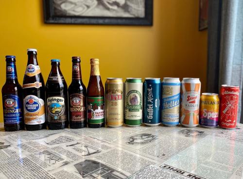 A lineup of various beer bottles and cans on a table with newspaper print, set against a yellow wall.