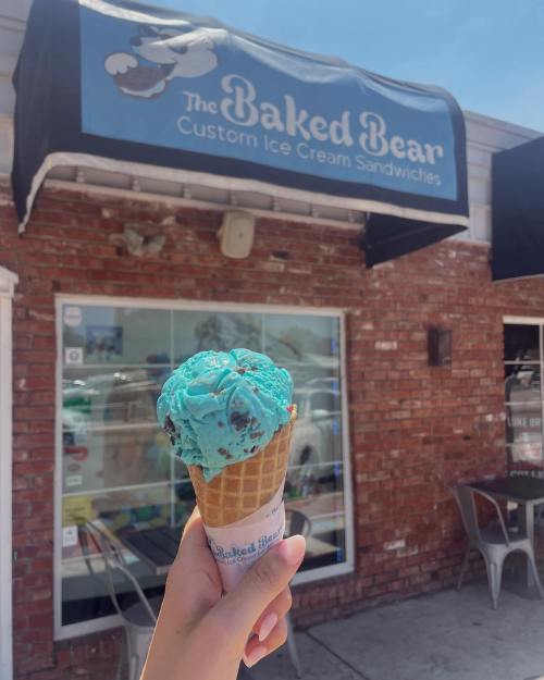 A hand holds a cone of blue mint chocolate chip ice cream in front of The Baked Bear ice cream shop.
