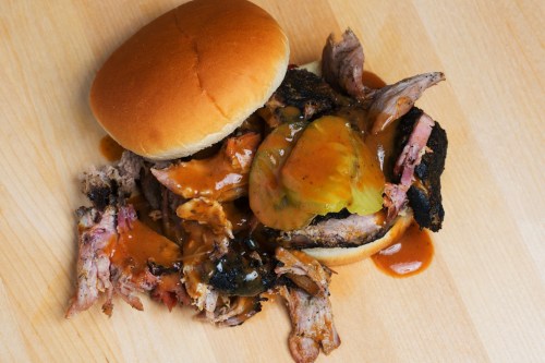 A messy pulled pork sandwich with pickles and barbecue sauce, spilling onto a wooden surface.