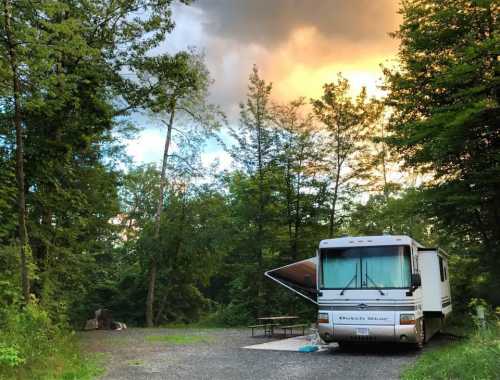 A parked RV in a wooded campsite, with trees surrounding and a colorful sunset in the background.