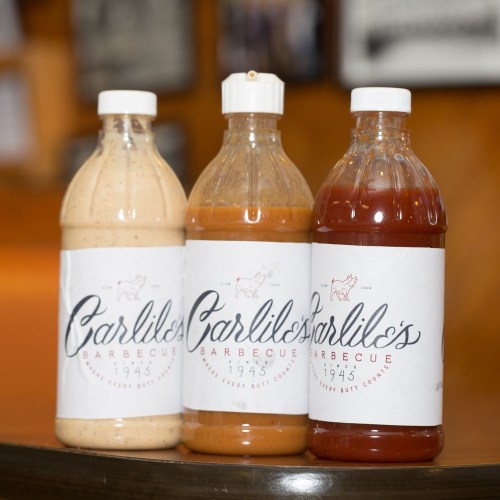 Three bottles of Carlile's Barbecue sauces, labeled with the year 1945, displayed on a wooden surface.