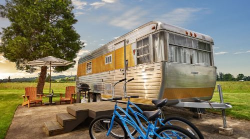 A vintage silver trailer with wooden accents, surrounded by a grassy area, patio furniture, and two blue bicycles.