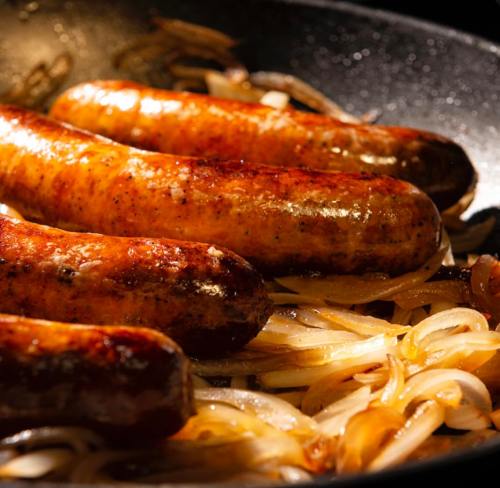 Sizzling sausages cooking with caramelized onions in a dark frying pan.