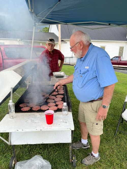 Two men grilling burgers at an outdoor event, with smoke rising and a red cup on the table.