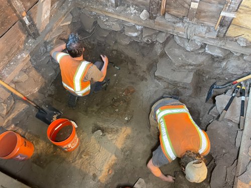 Two workers in safety vests excavate a dirt pit, surrounded by tools and buckets, in a wooden structure.