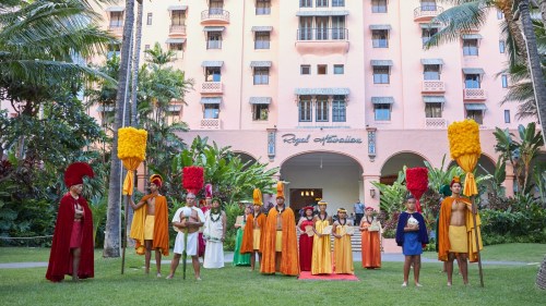 A group of people in colorful traditional costumes stands in front of a tropical hotel, surrounded by lush greenery.