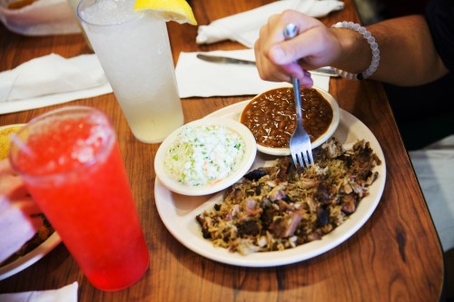 A plate of pulled pork with coleslaw and baked beans, accompanied by two colorful drinks on a wooden table.