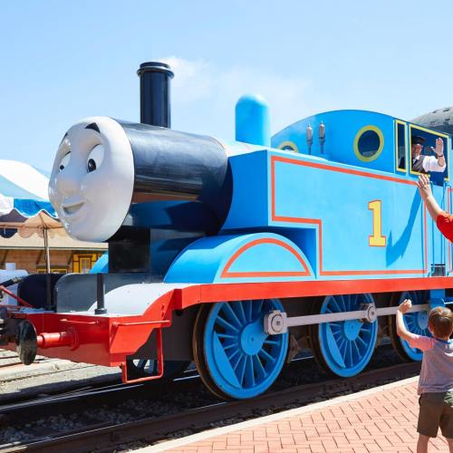 A large blue train with a smiling face, with a child waving beside it at a sunny outdoor event.