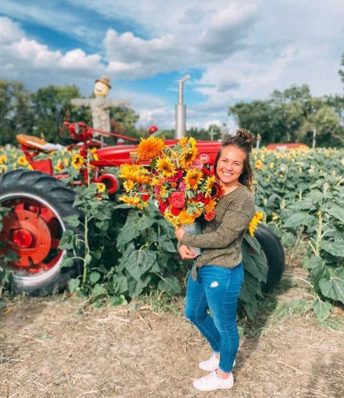 A smiling woman holds a bouquet of sunflowers in a field, with a red tractor and blue sky in the background.