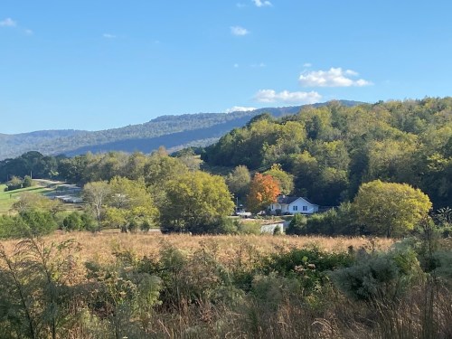 A scenic view of a house surrounded by trees and hills, with a clear blue sky and a hint of autumn colors.