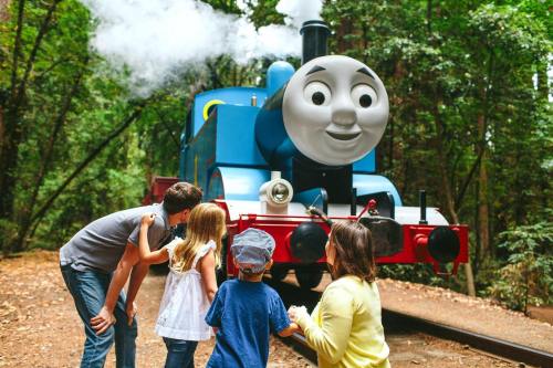 A cheerful blue train with a smiling face is surrounded by four children in a forest setting.