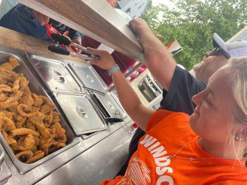 A young woman in an orange shirt uses tongs to serve food from a counter while a man assists her.