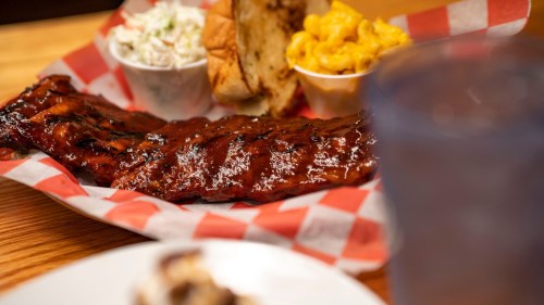 A plate of barbecue ribs with coleslaw, macaroni and cheese, and a slice of bread, served on a checkered paper.