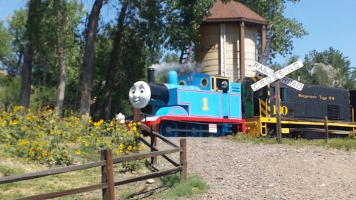 A blue train with a smiling face near a water tower and a railroad crossing sign, surrounded by sunflowers.