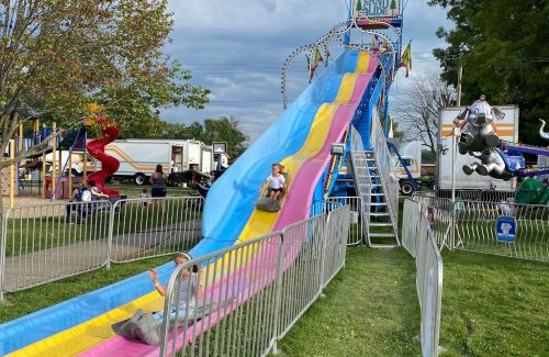 Two children slide down a colorful slide at a fair, with rides and attractions in the background.