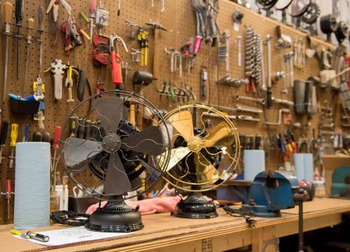 Two vintage fans on a workbench, surrounded by various tools and equipment on a pegboard wall.
